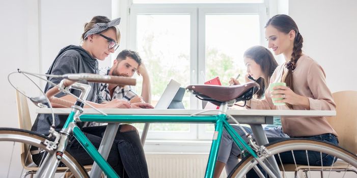 Four proficient freelancers and independent contractors co-working on tablets and laptops, connected to internet at a shared desk behind a commuter bike in a modern work hub