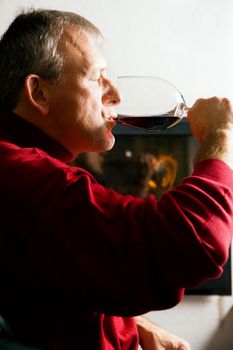 Mature man drinking a glass of read wine in front of his fireplace