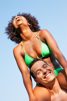 Couple in love - Woman of color in bikini sitting on her man’s shoulders under blue sky - summer and fun