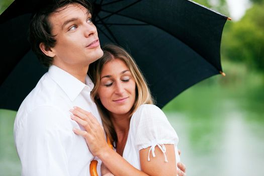 Couple (man and woman) at a lake in summer rain with an umbrella, he is sheltering her from the drops, holding his girl in his arms