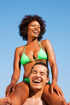 Couple in love - Woman of color in bikini sitting on her man’s shoulders under blue sky - summer and fun