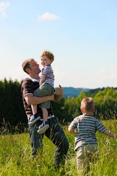 Father with two little boys playing in the grass on a summer meadow carrying one of the kids