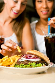 Two women - one is African American - eating hamburger and drinking soda in a fast food diner; focus on the meal