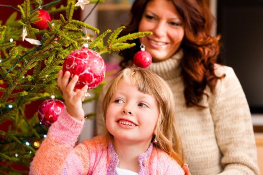 Young girl helping her mother decorating the Christmas tree, holding some Christmas baubles in her hand