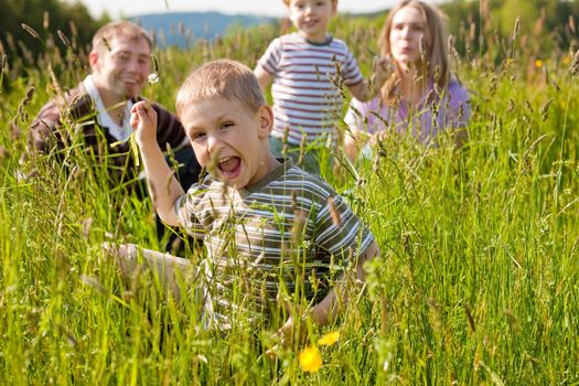 Very happy family with two kids sitting in a  meadow in the summer sun in front of a forest and hills, they are nearly hidden by the high grass, on boy is running towards the viewer