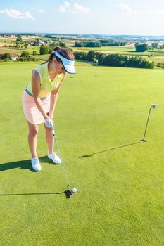 Full length of an attractive fit woman smiling while holding a putter golf club before hitting the ball during professional practice on putting green