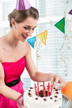 Portrait of a beautiful woman smiling while putting red candles on a birthday cake for her anniversary indoors in a decorated room