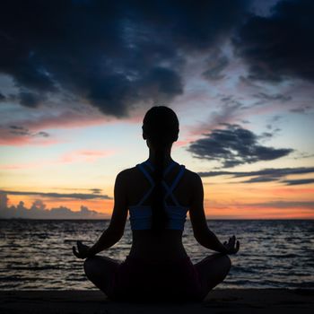 Full length rear view of a fit woman sitting in lotus position while practicing yoga on the beach at twilight during summer vacation in Flores Island, Indonesia