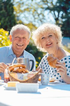 Happy elderly woman and man eating breakfast sitting in their garden outdoors in summer, eating bread rolls and drinking coffee