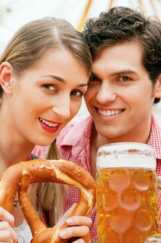 Couple in traditional German costume in a beer tent, he is having a drink, she a pretzel, scene could be located at the Oktoberfest or any Dult