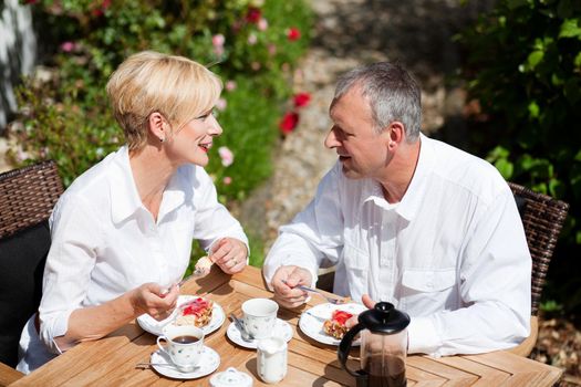 Mature or senior couple having coffee and strawberry cake on the porch in front of their home, it is summer and the roses look beautiful