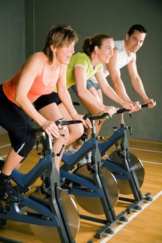 Three people cycling in a gym or fitness club, dressed in colorful clothes; focus on the mature woman in front