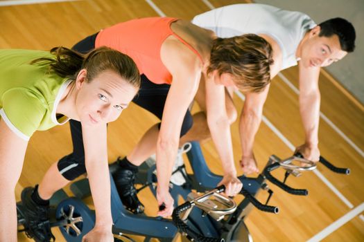 Three people cycling in a gym or fitness club, dressed in colorful clothes; focus on the girl in green looking into the camera