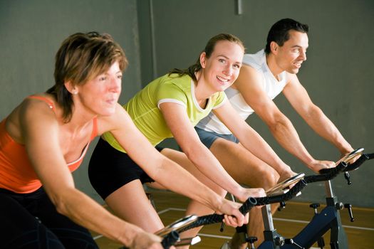 Three people cycling in a gym or fitness club, dressed in colorful clothes; focus on girl in the middle