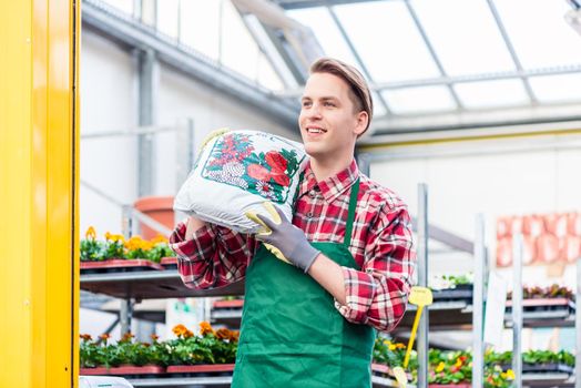 Portrait of a cheerful young man carrying a bag of high-quality potting soil while working as floristry specialist in a modern flower shop