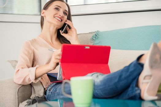 Successful female entrepreneur or independent contractor talking on mobile phone while sitting on sofa during remote work on tablet in a modern co-working space