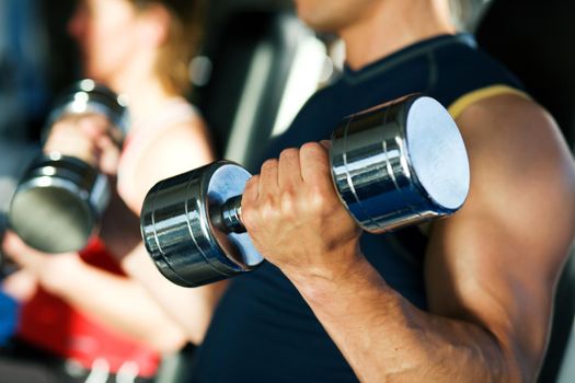 Strong man exercising with dumbbells in a gym, in the background a woman also lifting weights; focus on hand and dumbbell