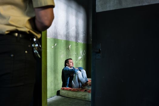 Side view of a young male prisoner thinking while sitting alone on a dirty mattress in an obsolete prison cell guarded by a police officer