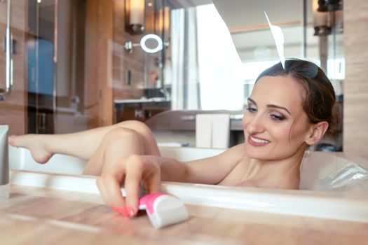 Woman having bath in hotel bathtub grabbing shaver for hair removal on her legs