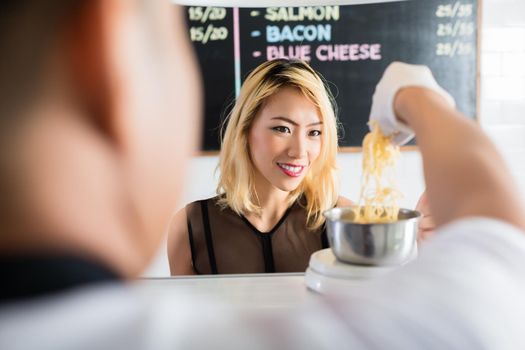 Asian woman watching intently as grated cheese is placed on a scale for weighing in a restaurant or deli in an over the shoulder view from behind the shop assistant