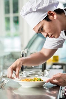 Handsome young Asian chef plating up food in a commercial kitchen carefully wiping around the dish before serving the customer