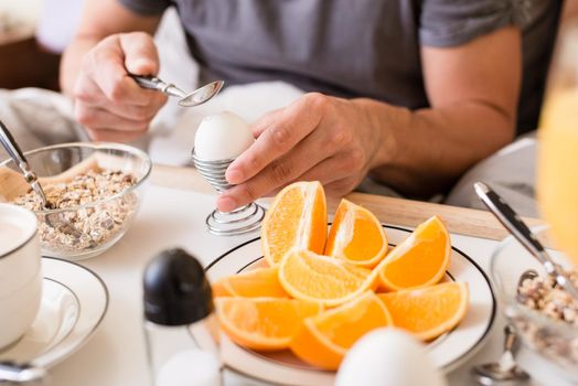Man cracking open a boiled egg for breakfast using a spoon with cereal and fresh oranges on the table in a healthy diet and lifestyle concept