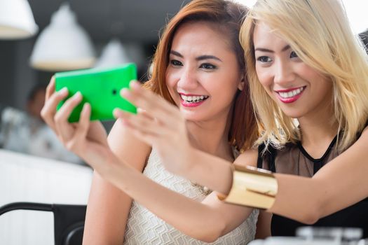 Two elegant Asian women posing for a selfie together on a mobile phone as they sit enjoying drinks in a restaurant