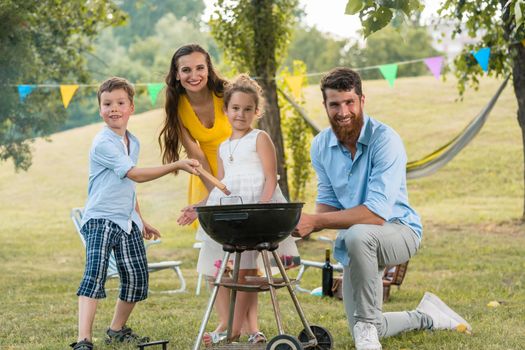 Portrait of a happy family with two children wearing summer casual outfits while posing together outdoors behind a round charcoal barbecue grill
