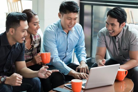 Two cheerful Chinese young men using a laptop while sharing business ideas with their colleagues during work in a successful multinational company