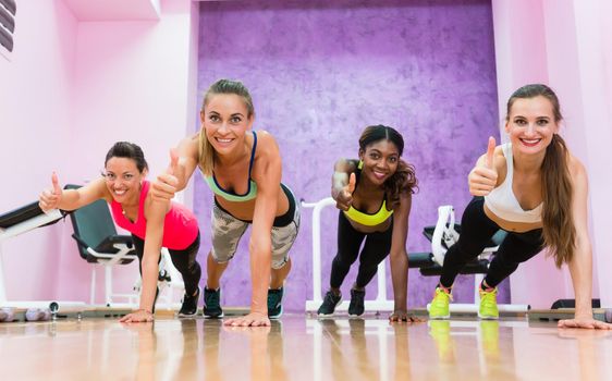 Group portrait of four happy women showing thumbs up from basic plank position, as a like sign for an efficient workout class in a trendy fitness club for ladies only