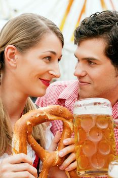 Couple in traditional German costume in a beer tent, he is having a drink, she a pretzel, scene could be located at the Oktoberfest or any Dult