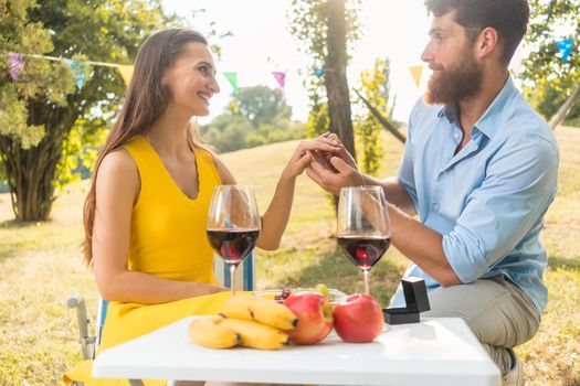 Young handsome man sitting on bended knee while showing to his beautiful girlfriend an engagement ring during romantic picnic with red wine and fresh fruits