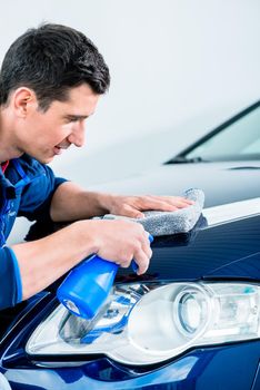 Man cleaning his luxury blue sedan car with a sponge and spray bottle as he works around the front headlamp