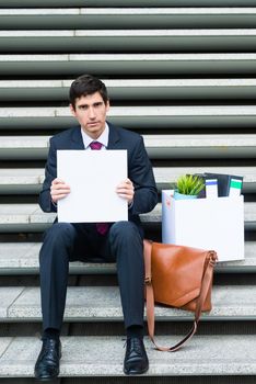 Worried unemployed businessman sitting on a flight of concrete steps in town with his office contents and blank sign seeking employment