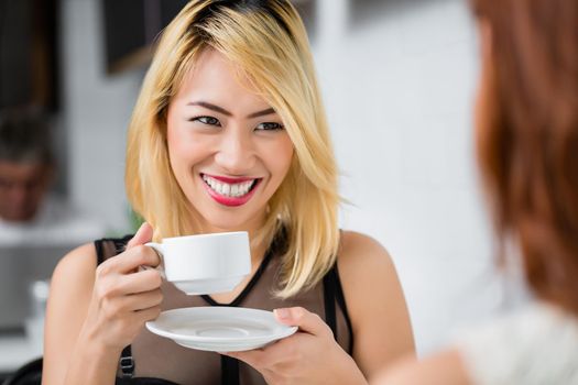 Beautiful blond Asian woman drinking coffee and smiling at her friend as they enjoy refreshments in a cafeteria, close up over the shoulder view of her face