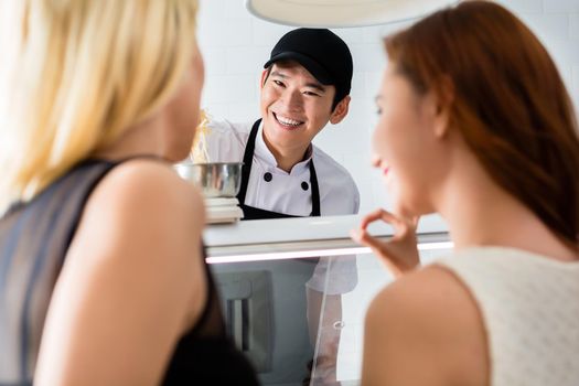 Smiling young man serving two ladies in a delicatessen weighing cheese in a counter top scale as they make their choices