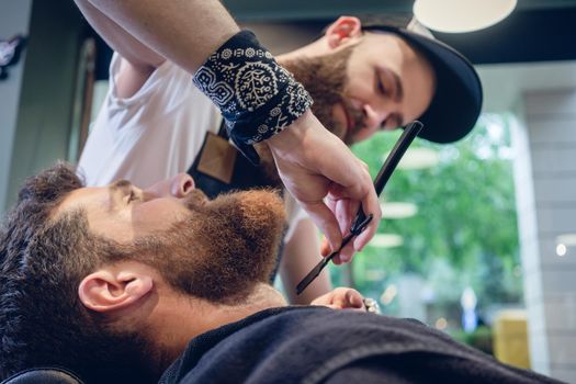 Side view headshot of a redhead bearded young man smiling, ready for shaving in the hair salon of a skilled barber with a classic straight razor in his hand