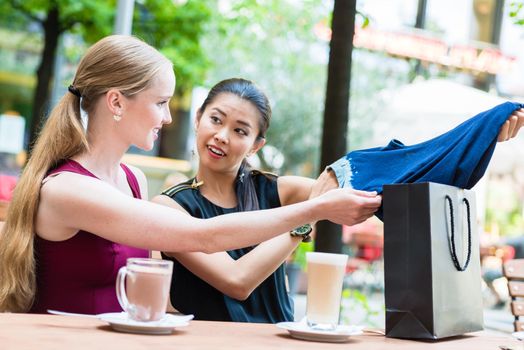 Pretty Asian woman showing a friend her purchase of a blue garment as they sit at a restaurant table enjoying refreshments