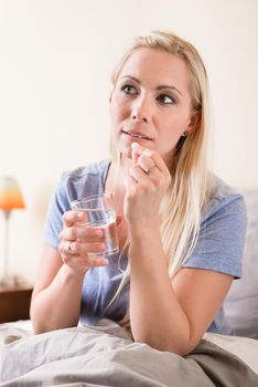 Young woman sitting in bed holding a glass of water taking a tablet while looking to the side in a healthcare concept