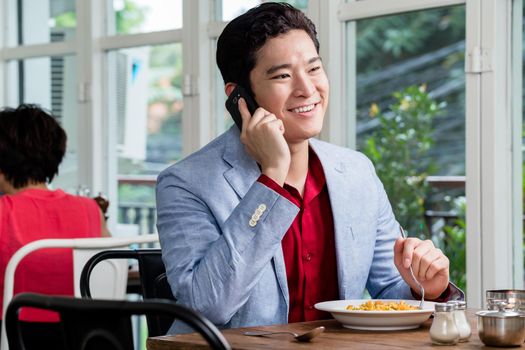 Asian businessman talking on his mobile phone with a smile as he relaxes in a restaurant enjoying a plate of food