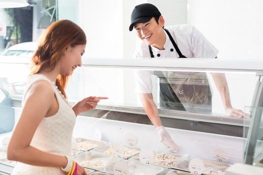 Smiling young Asian worker assisting a customer in a delicatessen as she selects food from a glass counter in the store