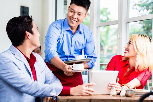 Friendly Asian waiter serving a stylish young couple coffee to accompany their meal in a restaurant with window background