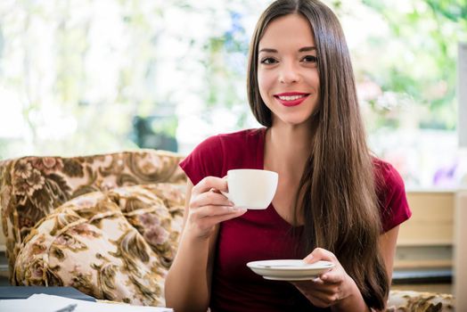 Pretty woman with a lovely smile relaxing on an armchair at home drinking coffee or tea from a handheld cup and saucer