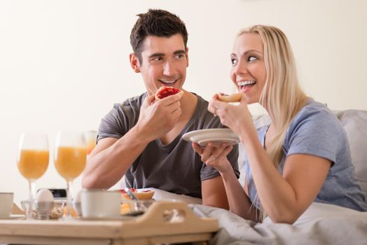 Happy playful couple enjoying breakfast in bed grinning at each other as they prepare to bite into toast and jam with a spread of orange juice, coffee and eggs on a tray in front of them