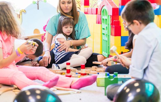 Dedicated young female kindergarten teacher holding a pre-school shy girl while watching children during playtime with wooden toy blocks in the classroom