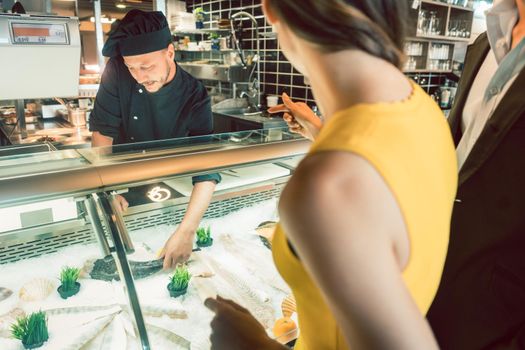 Experienced master chef taking a fresh fish from the freezer to cook it for a female customer and her partner at a trendy restaurant