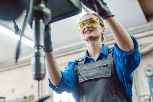 Woman worker in metal workshop using pedestal drill to work on piece