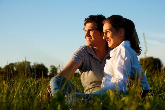 Young couple sitting together on a green meadow in summer looking into a golden sunset contemplating and daydreaming their future (marriage, children, divorce, eventual death and so on)