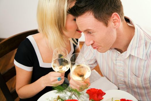 Young couple romantic dinner: both holding white whine glasses, she is whispering something; focus on his face