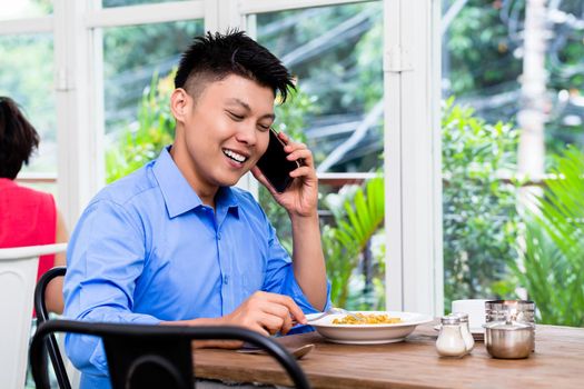 Young Asian man chatting on his mobile phone as he sits at a table in a restaurant with a plate of food in front of him smiling as he listens to the conversation